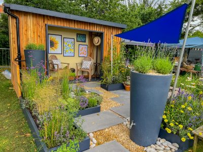 Designer Garden by Sally Leaney titled Adapting to Change. it has a small, covered seating area at the back and contains square planters around the edge. The garden has grey slabs in a pattern leading you through the garden and the rest of the open space is covered with small beige pebbles. There are lots of options for conserving water and providing shade for plants.