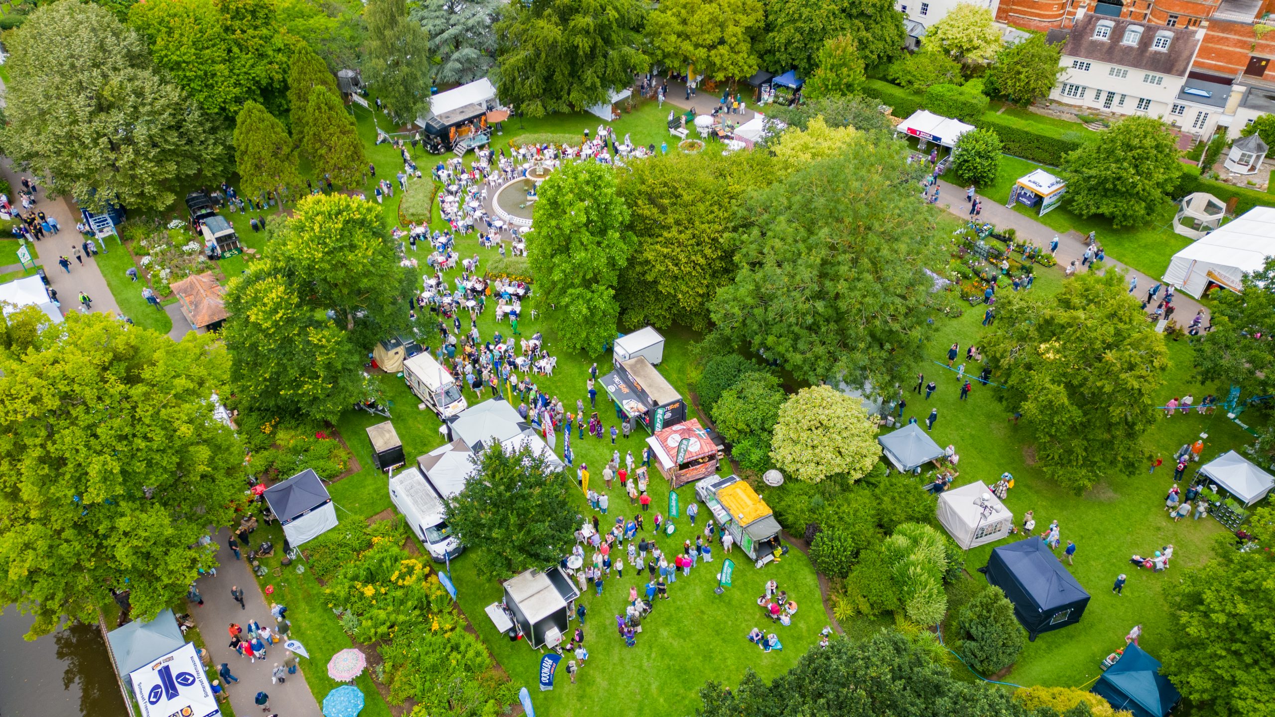 Drone view of the fountain area of Vivary Park with people milling about the catering area and views of the plant village and the bees and honey tent.